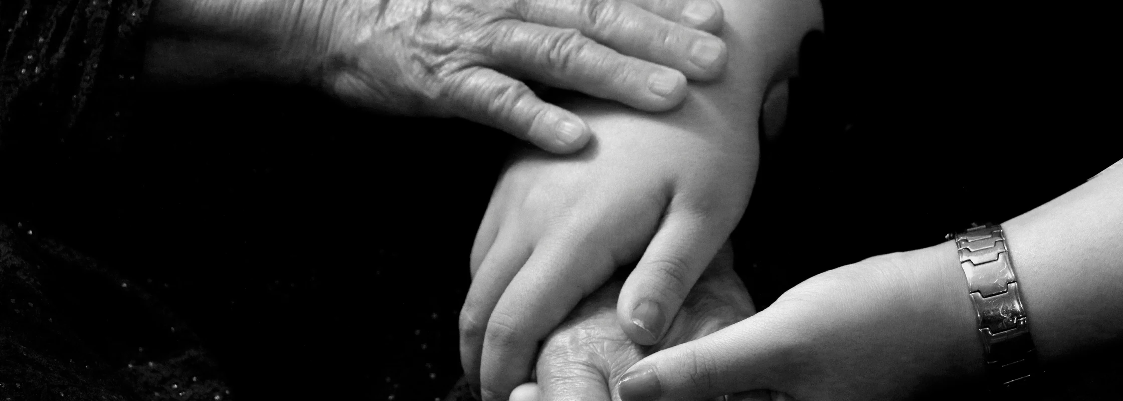 Three generations of hands clasped together — an elderly hand, a child's hand, and an adult hand